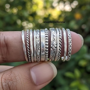 Op de afbeelding: Een verzameling zilveren ringen met verschillende ontwerpen, waaronder gedraaide, patroon- en gladde banden. De ringen zijn op een vinger gestapeld en tonen de verschillende texturen en stijlen. De achtergrond is een wazig groen.