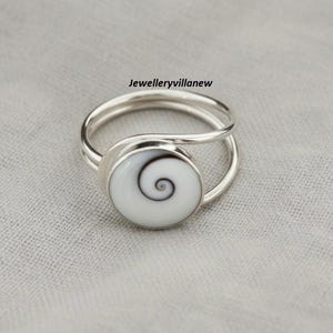 May include: A silver ring with a white, spiral-patterned stone. The ring has a double band design and the text "Jewelleryvillanew" is visible in the image. The stone is circular and features a dark brown spiral design.