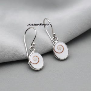 Peut inclure: Boucles d'oreilles en argent avec des pendentifs ovales blancs ornés d'un motif en spirale marron. Les boucles d'oreilles ont une fermeture à crochet. Le texte "Jewelleryvillanew" est visible.