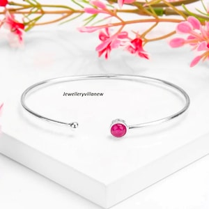 Könnte beinhalten: Ein silbernes Armreif-Armband mit einem einzigen rosafarbenen Edelstein. Das Armband ist offen und hat an einem Ende eine kleine Kugel.