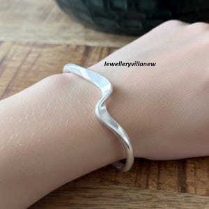 May include: A silver cuff bracelet with a flowing, wavy design. The bracelet is worn on a wrist. The text "Jewelleryvillanew" is visible on the image.