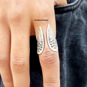Puede incluir: Un anillo plateado con forma de alas abiertas. El anillo se lleva en un dedo y presenta patrones de plumas detallados. El dise&ntilde;o del anillo es sim&eacute;trico, con cada ala reflejando la otra. El anillo es una joya.