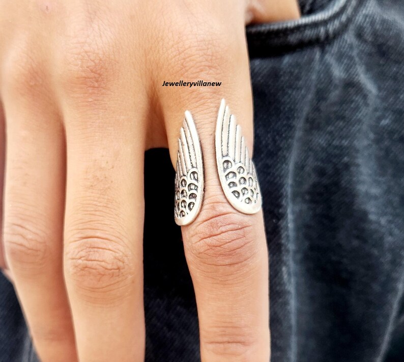 Puede incluir: Un anillo plateado con forma de alas abiertas. El anillo se lleva en un dedo y presenta patrones de plumas detallados. El dise&ntilde;o del anillo es sim&eacute;trico, con cada ala reflejando la otra. El anillo es una joya.