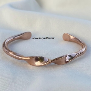 May include: A rose gold-colored cuff bracelet with a twisted design. The open-ended bracelet is made of a shiny metal and features the text "Jewelleryvillanew". The bracelet is displayed on a white surface.