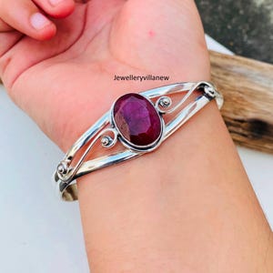 Könnte beinhalten: Ein silbernes Armreif-Armband mit einem großen, ovalen Rubin-Edelstein, der in der Mitte eingesetzt ist. Das Armband hat ein zartes, wirbelndes Design.