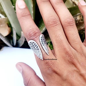 Puede incluir: Un anillo plateado con forma de alas de &aacute;ngel, usado en un dedo. El anillo tiene patrones detallados de plumas y se envuelve alrededor del dedo. El anillo se muestra sobre un fondo de follaje verde.