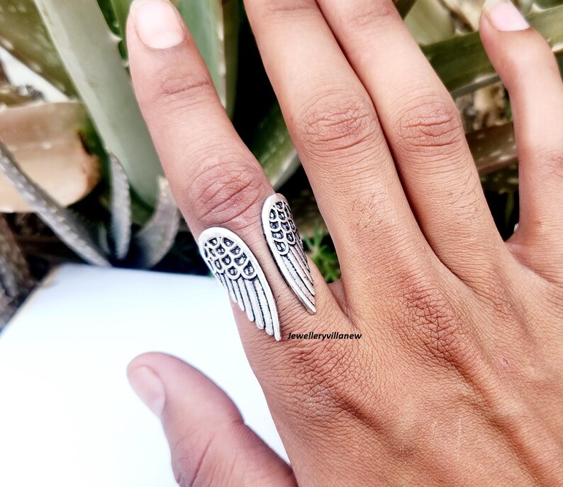 Puede incluir: Un anillo plateado con forma de alas de &aacute;ngel, usado en un dedo. El anillo tiene patrones detallados de plumas y se envuelve alrededor del dedo. El anillo se muestra sobre un fondo de follaje verde.