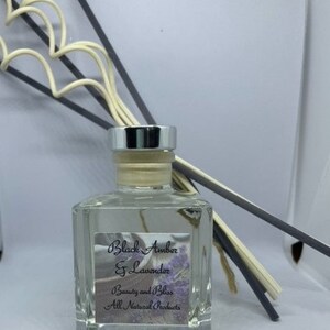 Peut inclure: Une bouteille en verre transparente avec un couvercle argent&eacute; et une &eacute;tiquette qui indique "Black Amber & Lavender" et "Beauty and Bliss All Natural Products". La bouteille est remplie d'un liquide clair et contient quelques b&acirc;tonnets.