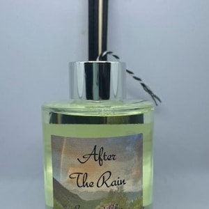 May include: A clear glass bottle with a silver lid and black and white reeds. The bottle contains a yellow liquid and has a label that reads "After The Rain" with a rainbow and a landscape scene. The label also says "Beauty and Bliss All Natural Products".