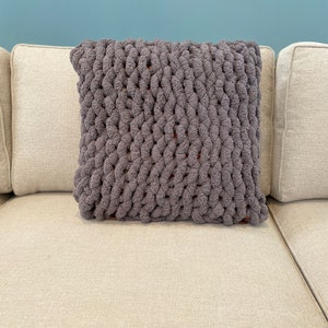 May include: A gray knitted throw pillow with a chunky knit texture. The pillow is on a light beige couch.