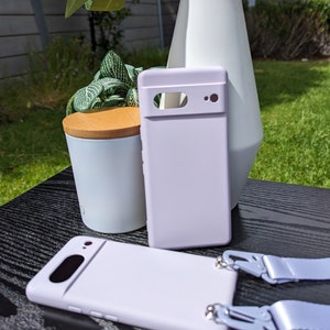 Pu&ograve; includere: Una custodia per telefono color lavanda con un ritaglio per la fotocamera e un laccio in tinta. La custodia &egrave; appoggiata su una superficie nera.