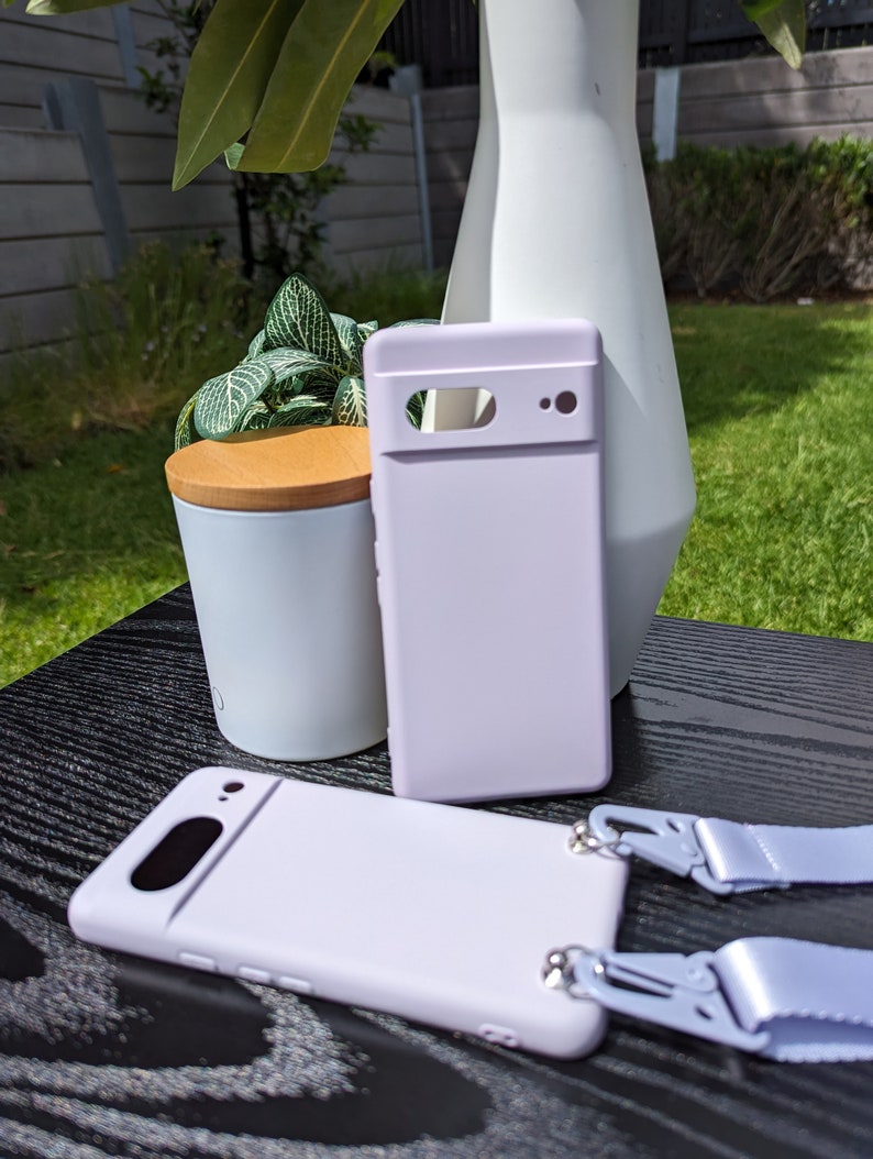 Pu&ograve; includere: Una custodia per telefono color lavanda con un ritaglio per la fotocamera e un laccio in tinta. La custodia &egrave; appoggiata su una superficie nera.