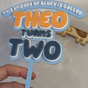 Op de afbeelding: Blauwe en witte taarttopper met de tekst "This episode of Bluey is called Theo turns two".