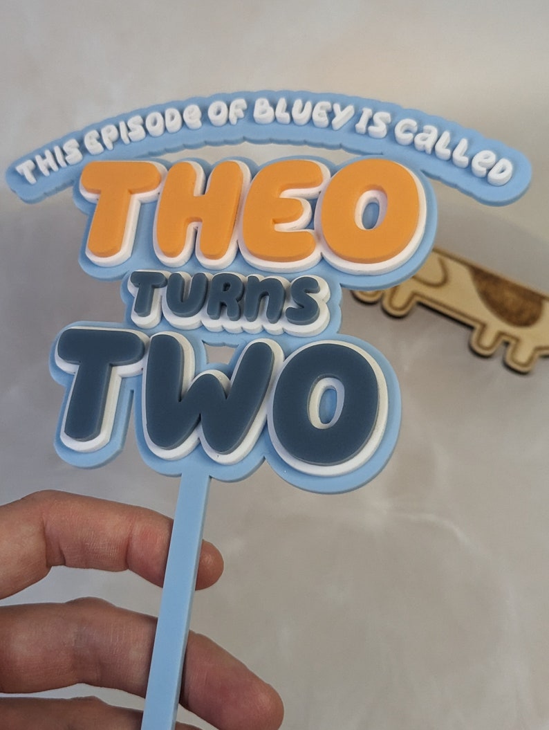 Op de afbeelding: Blauwe en witte taarttopper met de tekst "This episode of Bluey is called Theo turns two".