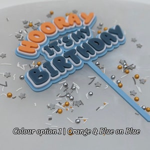Op de afbeelding: Een blauwe en oranje taarttopper met de woorden "Hooray It's My Birthday" in een vet, 3D-lettertype. De topper staat op een witte achtergrond met zilveren en gouden sprinkles.
