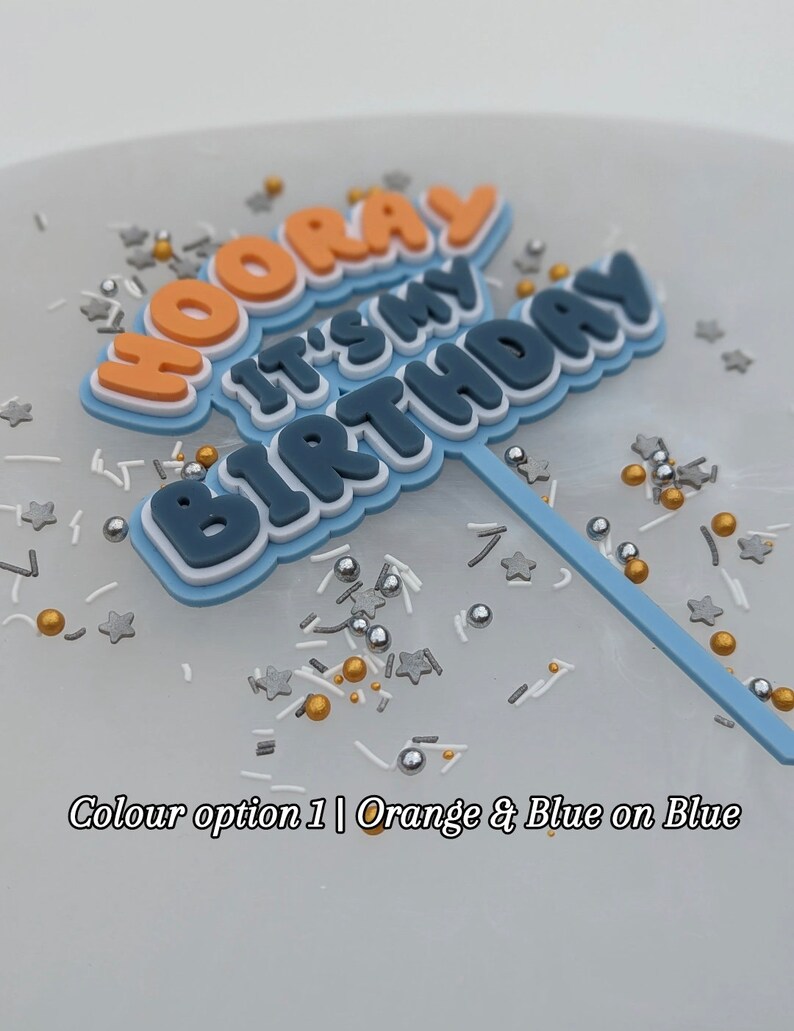 Op de afbeelding: Een blauwe en oranje taarttopper met de woorden "Hooray It's My Birthday" in een vet, 3D-lettertype. De topper staat op een witte achtergrond met zilveren en gouden sprinkles.
