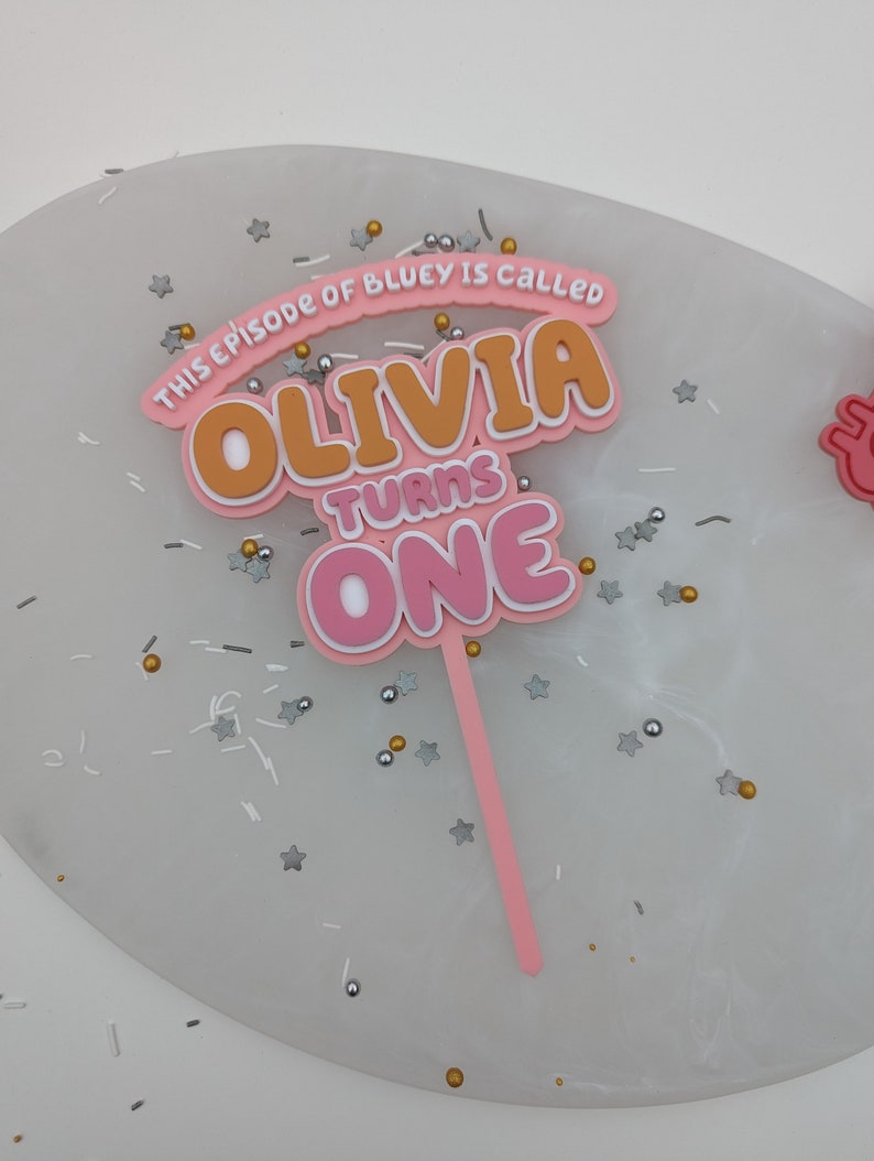 Op de afbeelding: Roze en oranje taarttopper met de tekst "This episode of Bluey is called Olivia turns one".