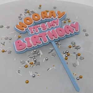 Op de afbeelding: Een taarttopper met de woorden "Hooray It's My Birthday" in oranje, roze en witte letters op een blauwe achtergrond. De topper staat op een wit oppervlak met gouden en zilveren sprinkles.