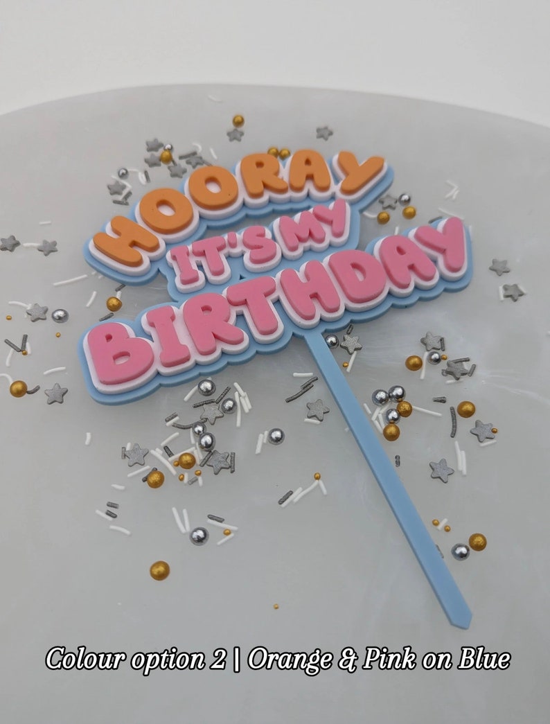 Op de afbeelding: Een taarttopper met de woorden "Hooray It's My Birthday" in oranje, roze en witte letters op een blauwe achtergrond. De topper staat op een wit oppervlak met gouden en zilveren sprinkles.