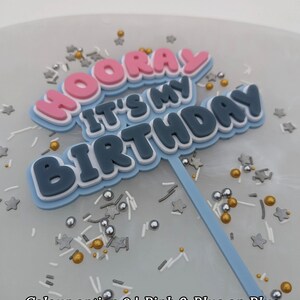 Op de afbeelding: Een blauwe taarttopper met de woorden "Hooray It's My Birthday" in roze, wit en blauwe letters. De topper staat op een blauwe achtergrond met gouden, zilveren en witte sprinkles.