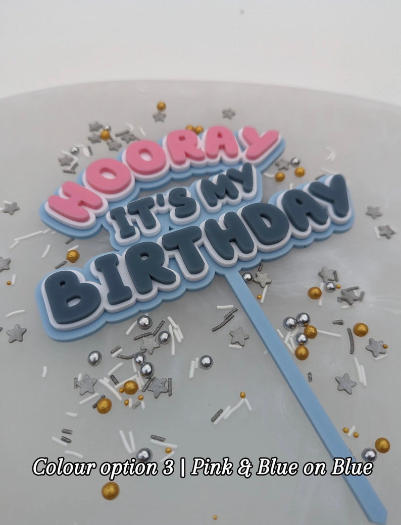 Op de afbeelding: Een blauwe taarttopper met de woorden "Hooray It's My Birthday" in roze, wit en blauwe letters. De topper staat op een blauwe achtergrond met gouden, zilveren en witte sprinkles.