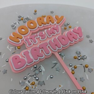 Op de afbeelding: Een roze en oranje taarttopper met de tekst "Hooray It's My Birthday" op een witte achtergrond met gouden en zilveren sprinkles.