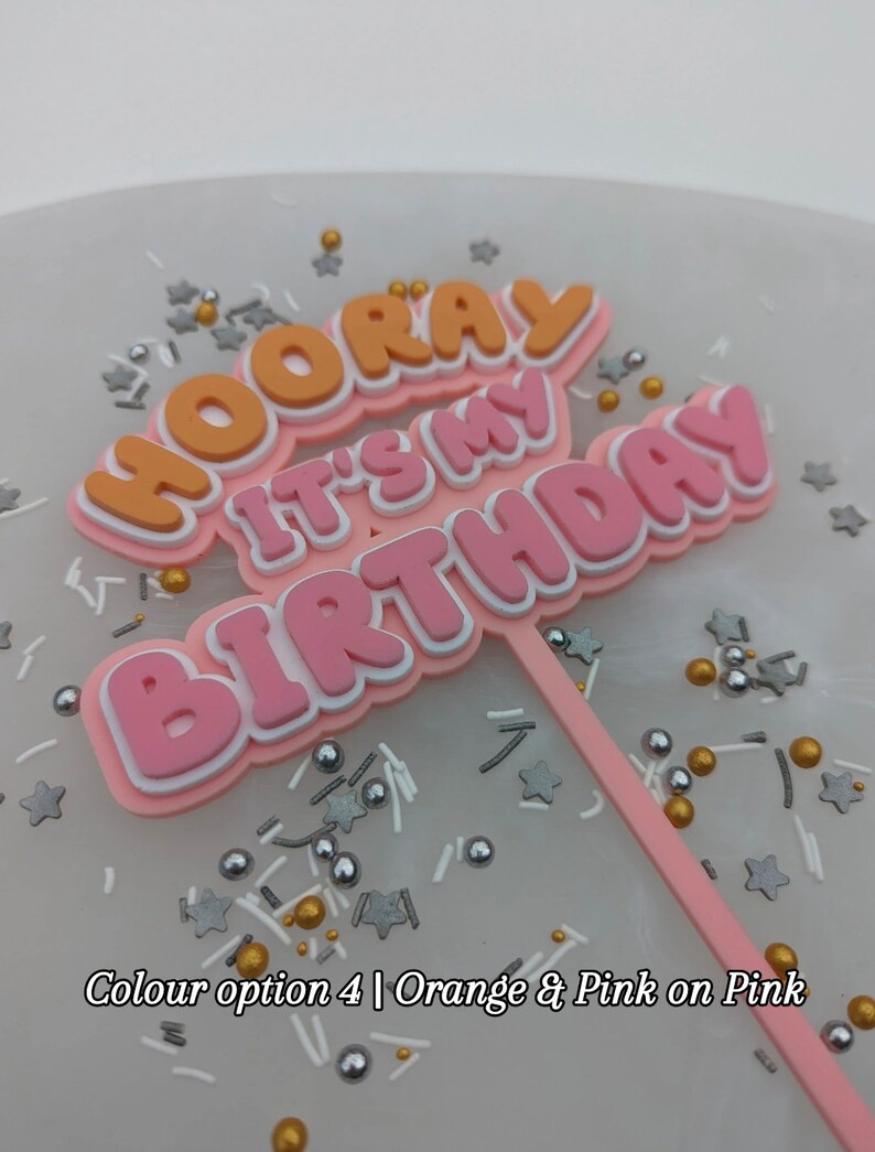 Op de afbeelding: Een roze en oranje taarttopper met de tekst "Hooray It's My Birthday" op een witte achtergrond met gouden en zilveren sprinkles.