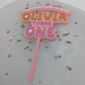 Puede incluir: Topper de pastel rosa y naranja con el texto "This episode of Bluey is called Olivia turns one".