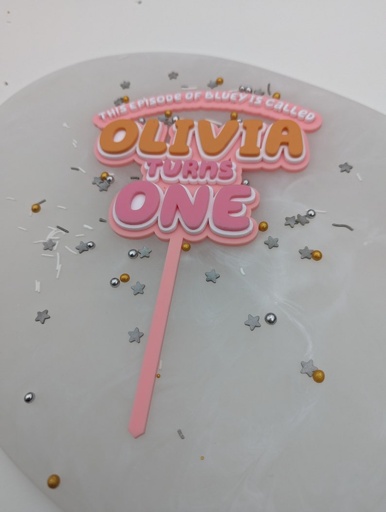 Puede incluir: Topper de pastel rosa y naranja con el texto "This episode of Bluey is called Olivia turns one".