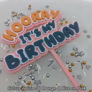 Op de afbeelding: Een roze taarttopper met de woorden "Hooray It's My Birthday" in oranje, blauw en witte letters. De topper staat op een witte achtergrond met gouden en zilveren sprinkles.