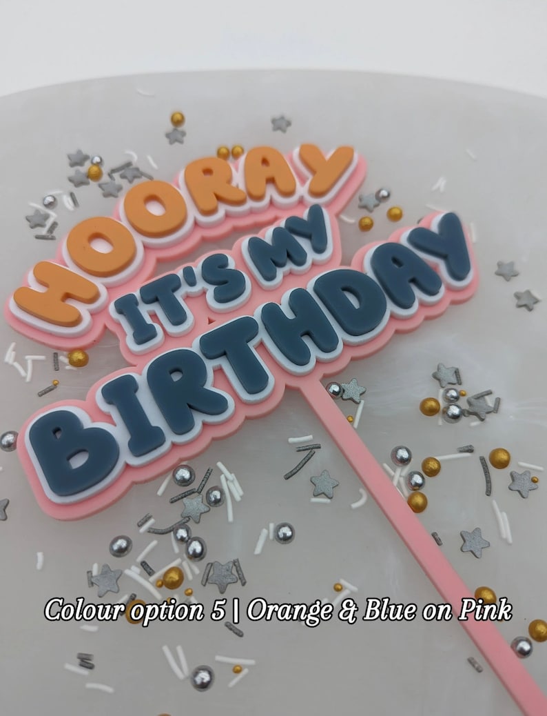 Op de afbeelding: Een roze taarttopper met de woorden "Hooray It's My Birthday" in oranje, blauw en witte letters. De topper staat op een witte achtergrond met gouden en zilveren sprinkles.