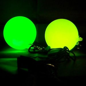 Può includere: Due globi luminosi, uno verde brillante e l'altro giallo-verde, sono il punto focale su uno sfondo scuro. Ogni globo ha un piccolo anello ed è collegato a un cordino scuro, suggerendo un portachiavi o un accessorio.