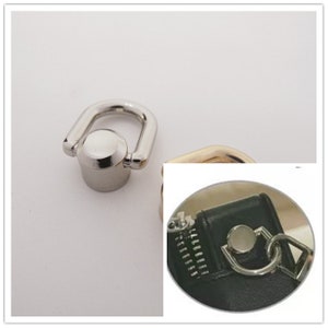 Può includere: Un anello a D in metallo argentato con una parte superiore arrotondata e una base cilindrica. L'anello a D è attaccato a un piccolo pezzo di metallo rotondo argentato. L'immagine mostra anche un primo piano di un oggetto nero con una cerniera e un anello a D.
