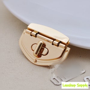 May include: A gold-coloured purse clasp with a rounded, shield-like shape. The clasp has a hinged design and a central locking mechanism. The metal hardware is shiny and reflective, with a smooth surface. The background is a neutral colour.