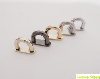 May include: A collection of five metal D-rings in various finishes: gold, black, gold, silver, and chrome. Each D-ring has a rectangular base and a curved top, suitable for bag hardware or craft projects.
