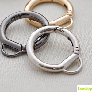May include: Close-up of three metal round spring clasp key rings in silver, gunmetal, and gold. The clasps are open, showing the spring mechanism. The key rings are on a light gray surface.