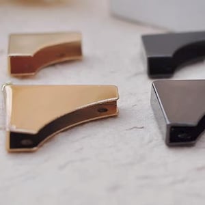 May include: Close-up of four metal corner brackets in gold and gunmetal gray. The brackets are L-shaped with a curved design. Small screws are scattered nearby. These hardware pieces are likely for crafting or furniture making.