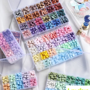 Pode incluir: Sortido de contas de cera coloridas em caixas organizadoras de plástico transparente. As contas são de várias cores, incluindo dourado, prateado, rosa, roxo, azul, verde e amarelo. Também são mostrados selos de cera, um carimbo e o texto "Luoshop".
