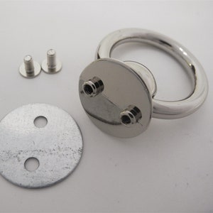 May include: A collection of silver metal hardware components. It includes a D-ring, two screws, and a circular plate with two holes. The D-ring is connected to a circular base with two screw holes.