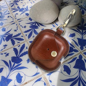 Peut inclure: Un étui en cuir marron avec un fermoir et un bouton dorés. L'étui est conçu pour un petit appareil électronique. Le fond présente un motif de carreaux floraux bleus et blancs.