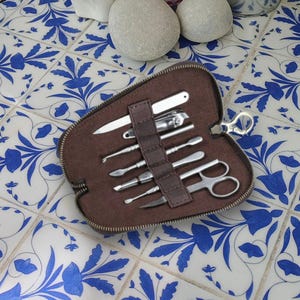 Könnte beinhalten: Ein braunes Maniküre-Set mit Reißverschluss und verschiedenen Edelstahlwerkzeugen, darunter Nagelknipser, Scheren und Nagelhautschieber. Das Set ist geöffnet und zeigt die Werkzeuge ordentlich angeordnet. Der Hintergrund ist eine weiße Fliese mit blauen Blumenmustern.