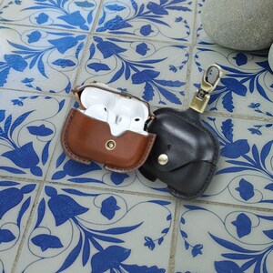 Könnte beinhalten: Zwei Leder-AirPod-Hüllen. Eine ist braun und offen und zeigt weiße Ohrhörer. Die andere ist schwarz und geschlossen, mit einem goldfarbenen Verschluss. Beide Hüllen haben einen Knopfverschluss. Die Hüllen liegen auf einer blau-weißen Fliesenoberfläche.