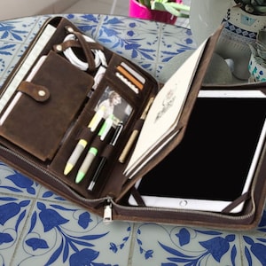 Könnte beinhalten: Ein braunes Leder-Zipper-Portfolio mit mehreren Fächern zum Tragen eines Tablets, Stiften und anderer Büroartikel. Das Portfolio ist geöffnet und das Tablet ist sichtbar.