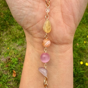May include: A gold-colored necklace with a sun pendant and a chain of colorful gemstones. The gemstones include yellow, orange, pink, and lavender stones. The necklace is held against a background of green grass.