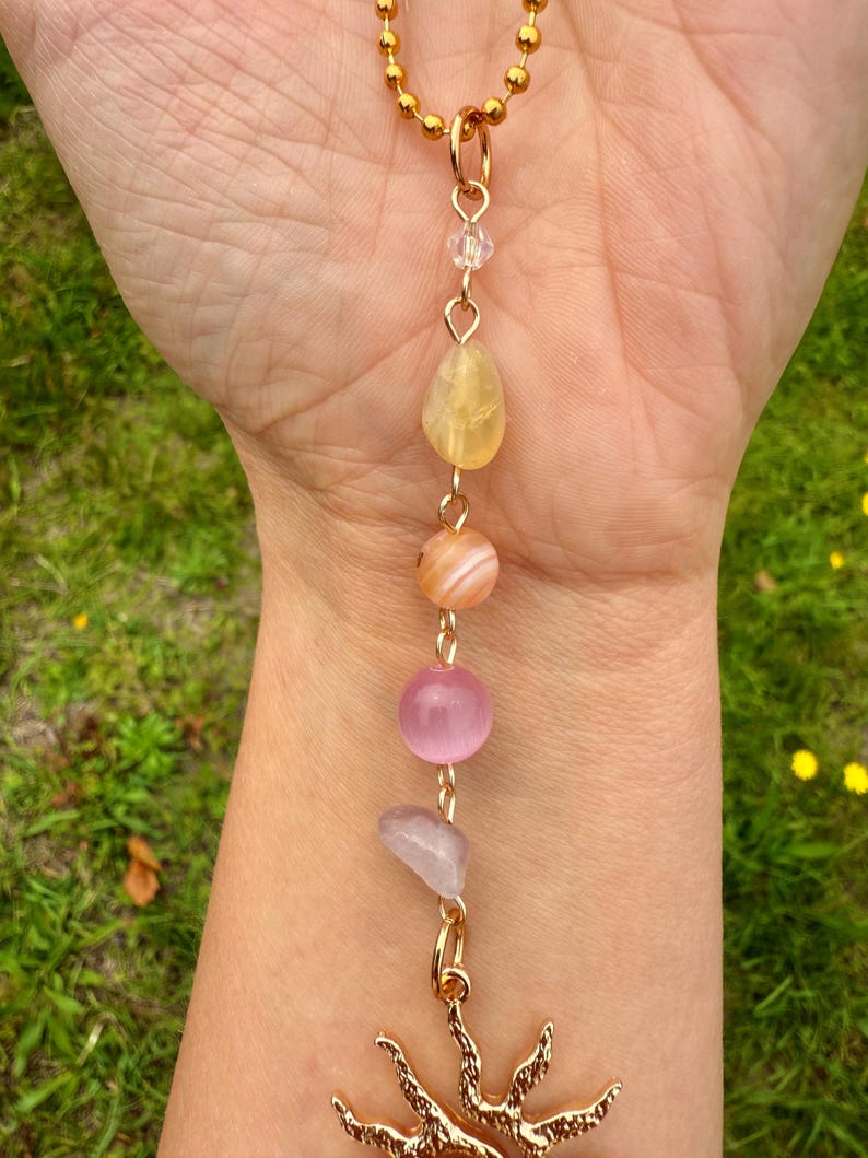 May include: A gold-colored necklace with a sun pendant and a chain of colorful gemstones. The gemstones include yellow, orange, pink, and lavender stones. The necklace is held against a background of green grass.