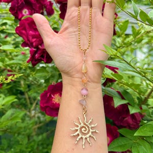 May include: A gold-colored necklace with a sun pendant and a chain of small beads. The necklace features a series of colorful gemstones in shades of yellow, orange, and pink. The sun pendant has a spiral center and radiating lines.