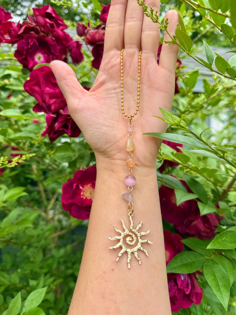 May include: A gold-colored necklace with a sun pendant and a chain of small beads. The necklace features a series of colorful gemstones in shades of yellow, orange, and pink. The sun pendant has a spiral center and radiating lines.