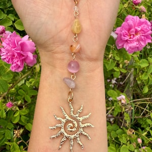 May include: Gold-colored necklace featuring a sun pendant and a strand of colorful gemstones. The pendant has a spiral center with radiating rays. The gemstones include yellow, orange, pink, and lavender beads. The necklace is displayed against a backdrop of pink roses.