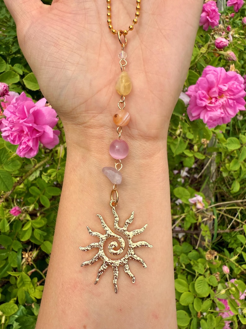May include: Gold-colored necklace featuring a sun pendant and a strand of colorful gemstones. The pendant has a spiral center with radiating rays. The gemstones include yellow, orange, pink, and lavender beads. The necklace is displayed against a backdrop of pink roses.