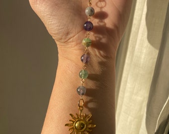 Puede incluir: Un collar de cadena dorada con un dije de sol y una variedad de cuentas de colores, incluyendo verde, morado y gris.